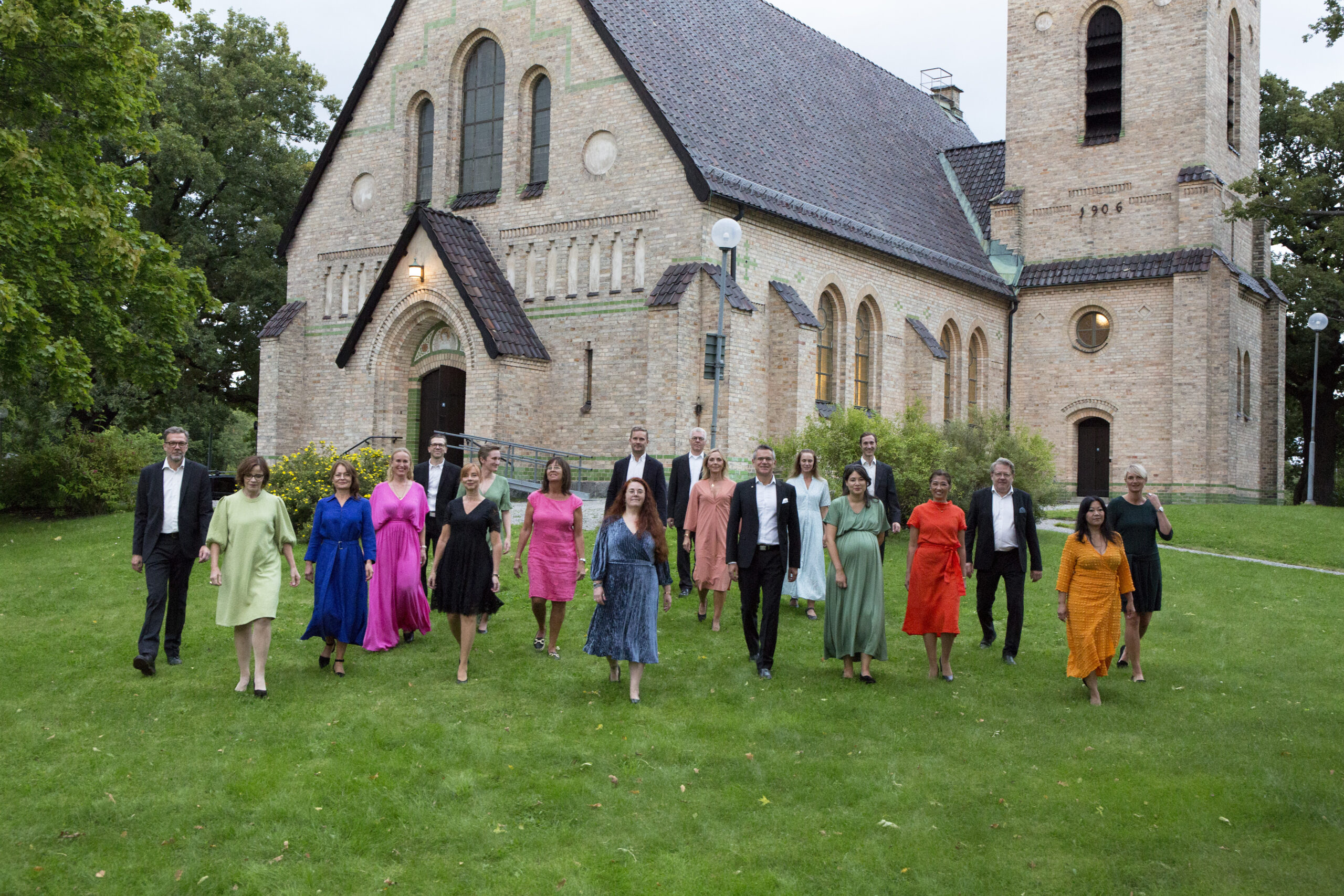 Gustavsbergs Vokalensemble i färgglada kläder i grönskan framför kyrkan